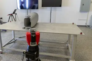 A lab worktable with a red and black device mounted underneath and a gray speaker-like object on top, with a TV screen on the wall behind.