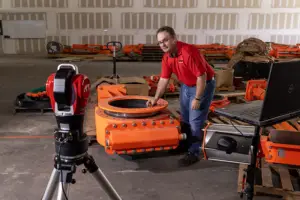A man in a red shirt inspects a large orange industrial component in a workshop, with a camera on a tripod and a computer monitor nearby.