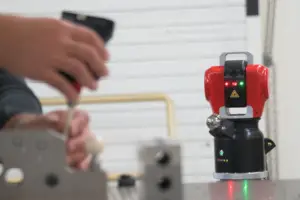 A small red and black robot sits on a table while a person in the background uses a smartphone.