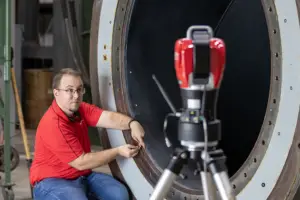 A man in a red shirt and jeans works on a large circular industrial hatch, with a tripod-mounted instrument in the foreground.