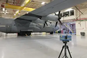 Large gray military transport aircraft inside a bright hangar, with a tripod-mounted device in the foreground.