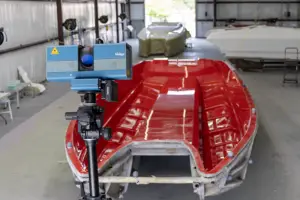 Red rigid-hull boat on a trailer in a workshop, with a blue measurement scanner mounted on a stand beside it.