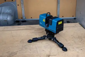 Blue laser scanner on a small tripod sits on a plywood floor in an industrial space.