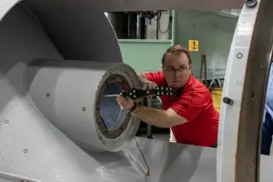 A man in a red shirt and safety gloves works on the circular opening of a large industrial machine in a factory.