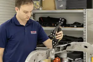 Man in a blue polo shirt works on a metal vehicle frame with a clamp tool in a workshop.