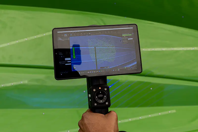 A hand holds a handheld device with a mounted screen displaying blue and green graphics against a bright green field background.