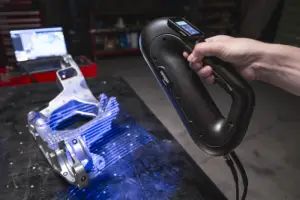 A person holds a black handheld device next to a metal mechanical part on a workshop bench with blue liquid spilled.