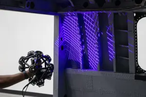 A hand holds a circular, spoked metal device in front of a high-tech lab rig illuminated by purple lights.