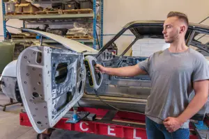A man in a gray T-shirt stands beside a disassembled car body on a trailer in a workshop.