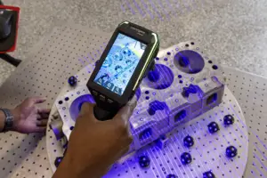 A person holds a handheld scanner over a circular perforated metal lab apparatus illuminated by purple light.