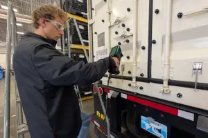 A worker wearing safety glasses and gloves works on the side of a large white truck trailer.