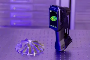 A blue-and-black handheld 3D scanner stands on a perforated metal surface beside a purple flower-shaped 3D-printed object.