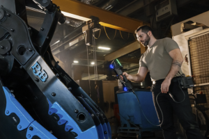 Technician using handheld 3D laser scanner to inspect large blue and black industrial machinery in a workshop.