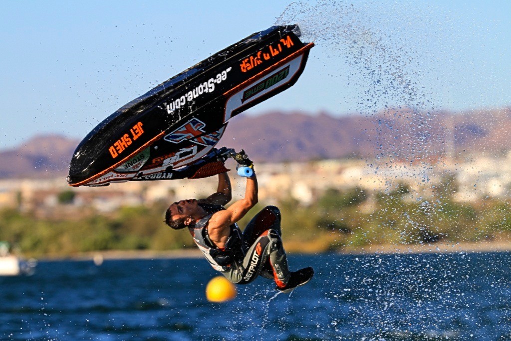 Professional jet ski rider performing an aerial backflip trick over water with mountains in the background